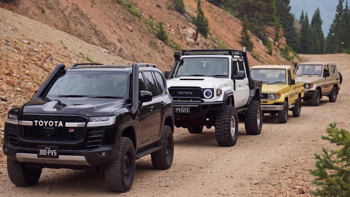 Four generations of Toyota LandCruiser on a mountain track: a black 300 Series, a white 79 Series dual cab, a yellow FJ75 ute and a sandy taupe FJ73
